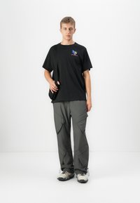 Joven de pie vistiendo una camiseta gráfica negra, pantalones cargo grises, zapatillas blancas y una bolsa de hombro negra frente a un fondo blanco.