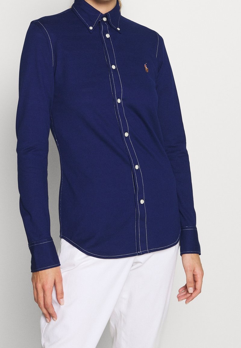 Woman wearing a fitted navy blue button-up shirt with white stitching details and white pants against a plain light background.