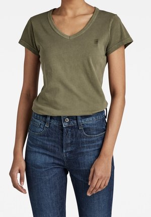 Žena v olivově zeleném tričku s výstřihem do V a krátkými rukávy, zastrčeném do tmavě modrých džínů s vysokým pasem, na bílém pozadí.