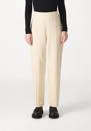 Pantalons taille haute couleur crème avec plis à l'avant, en tissu léger. Associés à des chaussures à lacets noires.