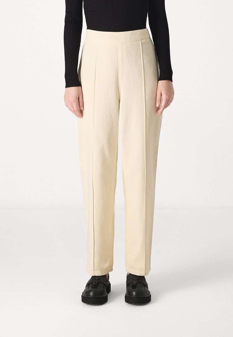 Pantaloni color crema a vita alta con pieghe frontali, realizzati in tessuto leggero. Abbinati a scarpe nere con lacci.