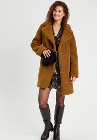 Manteau en fausse fourrure marron avec un col oversized, deux boutons et un tissu texturé. Le modèle porte une robe à motifs et des bottes noires montantes.