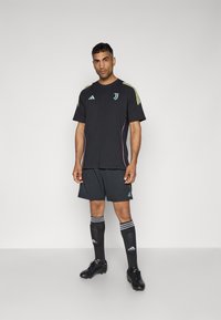 Schwarzes Sportoutfit mit einem kurzärmeligen Hemd mit Schulterstreifen, passenden Shorts, knielangen Socken und schwarzen Fußballschuhen.