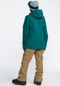 Veste imperméable bleu-vert avec une capuche, associée à un pantalon de neige beige et des bottes noires. Le tissu semble lisse, avec des coutures et des poches minimales.