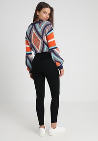 Kleurige gebreide trui met geometrische patronen, oranje en blauwe accenten, gecombineerd met hoge zwarte jeans en witte sneakers.