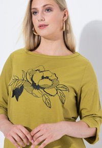 Top verde oliva con mangas de 3/4, que presenta un diseño gráfico de flores negras con detalles punteados y hojas, realizado en una tela suave y elástica.