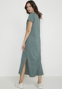 Long, teal cotton dress with short sleeves and side slits. Smooth texture, relaxed fit, falls below the knee, paired with light sneakers.