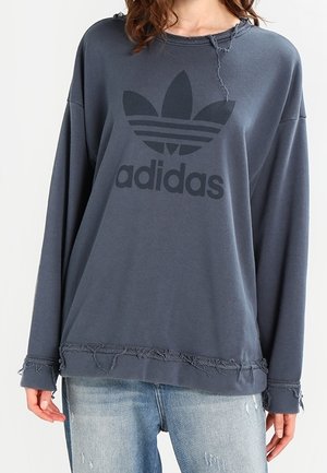 Kobieta ubrana w luźną, ciemnoszarą bluzę Adidas z postrzępionymi krawędziami oraz jasnoniebieskie, przetarte dżinsy na jednolitym tle.