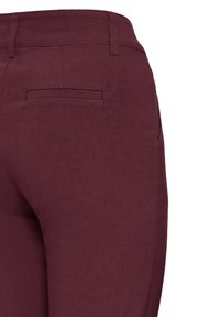 Gros plan sur une personne portant un pantalon bordeaux, montrant l’arrière de la taille avec une poche passepoilée et des passants de ceinture.