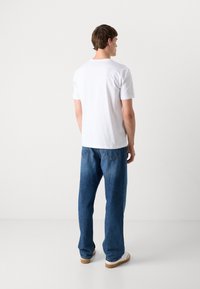 Witte kortekorte mouwen t-shirt gecombineerd met losse blauwe jeans; de schoenen van het model zijn wit met bruine zolen. Eenvoudige achtergrond.