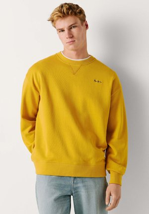 Joven con sudadera de cuello redondo color mostaza y vaqueros azul claro, de pie frente a un fondo blanco liso.