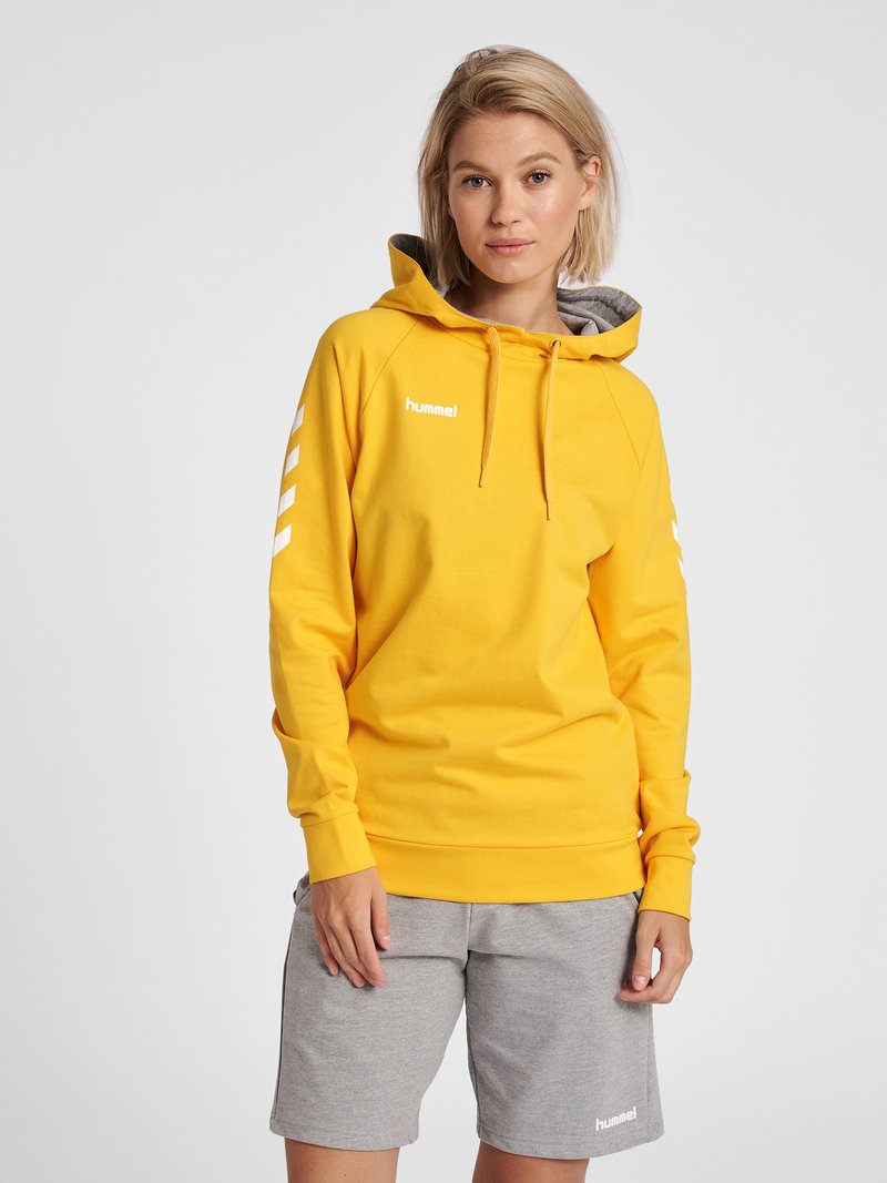 Junge Frau trägt einen gelben Hoodie mit weißen Chevron-Streifen und graue Shorts, sie steht vor einem schlichten hellen Hintergrund.