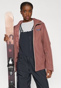 Casaco de esqui da Patagonia em rosa pó com capuz, bolsos com fecho; combinado com um macacão preto e uma camisa cinza. Segurando esquis com ligações pretas.