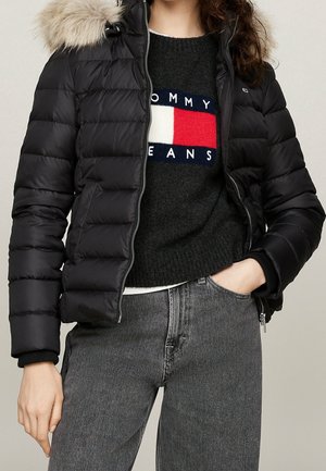 Mujer con chaqueta acolchada negra con capucha de piel sobre un suéter negro de Tommy Jeans y jeans grises, con una mano en el bolsillo.