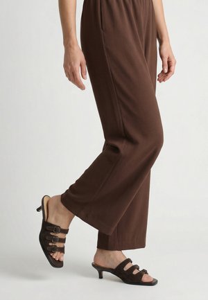 Frau trägt braune Hosen mit weitem Bein und dunkelbraune Sandalen mit Absatz und drei Schnallenriemen, steht auf weißem Hintergrund.