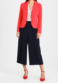 Blazer de coral, blusa blanca con lunares, pantalones de tiro ancho en azul marino y tacones rojos. Texturas suaves, diseño ajustado con botones y bolsillos.