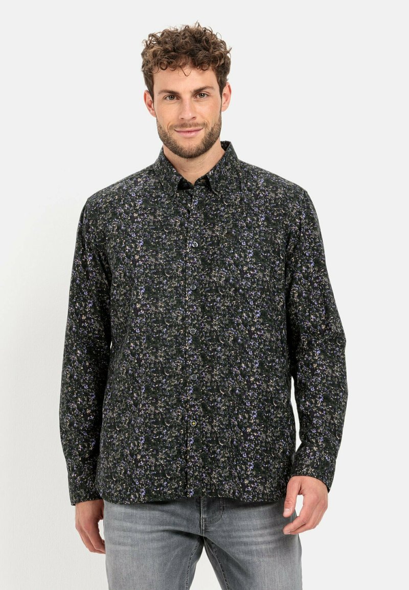 Ein Mann mit lockigem Haar trägt ein dunkelblaues, floral gemustertes Hemd und hellgraue Jeans, während er vor einem schlichten weißen Hintergrund steht.