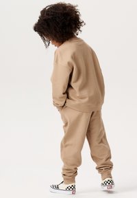 Beiger Sweater und passende Jogginghose aus weichem Material, mit lässigem Schnitt. Das Outfit wird mit schwarz-weißen Karo-Slippern kombiniert.
