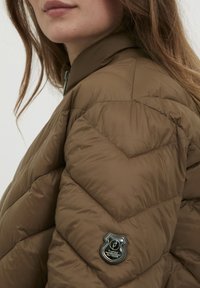 Hnedá puffa bunda s chevron vzorom, vysokým golierom a čiernou nášivkou s textom. Hladká, ľahká tkanina s lesklým dokončením.