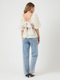 White floral lace blouse with a deep V-back and tied detail. Paired with light blue high-waisted jeans and black heeled mules.