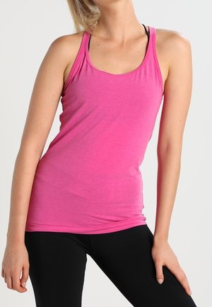 Nainen, jolla on kirkkaan pinkki hihaton paita ja mustat leggingsit, poseeraa yhdellä kädellä lanteillaan yksinkertaisen taustan edessä.