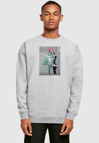 Grå sweatshirt med en tryckt design av en pojke som sträcker sig efter en stjärna på toppen av ett grönt träd med en röd stjärna, mot en texturerad bakgrund.