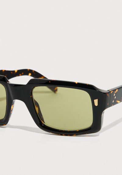 Μαύρα ορθογώνια γυαλιά ηλίου από χελώνα με φακούς με πράσινη απόχρωση και μικρό χρυσό έμβλημα ασπίδας στη γωνία του σκελετού.