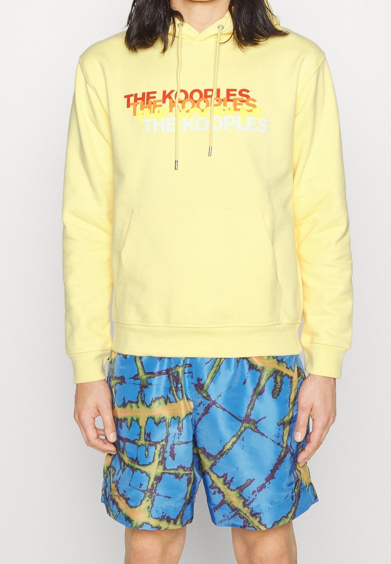 Personne portant un sweat à capuche jaune pâle avec le texte multicolore "THE KOOPLES" et un short bleu à motif abstrait, debout sur un fond uni.