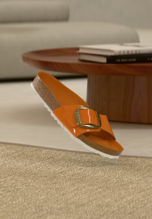 Oranž toonirse lamekinga korkpõhjaga, valge tekstuuriga välistallaga ja suure kullast pandlaga, mis asub puidust kohvilaua lähedal, millel on virnastatud raamatud.