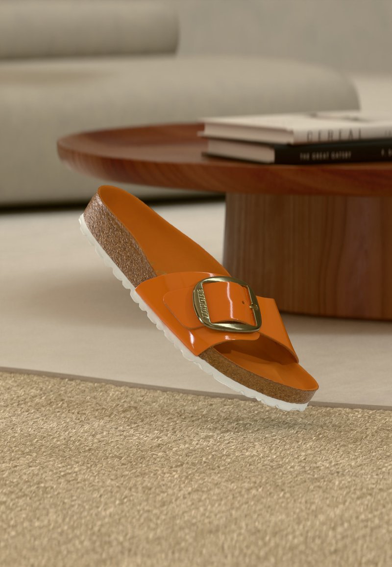 Πορτοκαλί σανδάλι με φτέρνα φελλού, λευκή υφασμάτινη εξωτερική σόλα και μεγάλο χρυσό κούμπωμα που βρίσκεται κοντά σε ξύλινο τραπεζάκι καφέ με στοίβες βιβλίων.