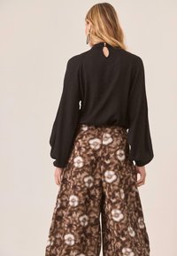 Blusa negra de manga larga con un detalle de abertura en la parte posterior, combinada con pantalones de pierna ancha que presentan un patrón abstracto en tonos marrón, crema y marrón claro.