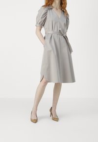 Vaaleanbeige mekko, jossa on V-neck, puffi hihat ja vyöllä varustettu vyötärö. Sivuhalkiot ja valuvan A-linjan siluetti. Beige korkokengät.