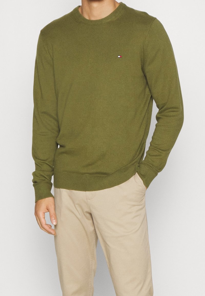 Homme portant un pull à col rond à manches longues de couleur vert olive avec un petit logo sur la poitrine et un pantalon beige, main dans la poche.