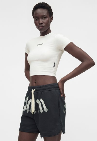 Λεπτή γυναίκα με κοντά κατσαρά μαλλιά φοράει λευκό κοντομάνικο τοπ και μαύρα σορτς με λευκό κορδόνι και μεγάλο κείμενο.