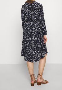 Granatowa sukienka w kwiatowy wzór, długie rękawy i wiązany w pasie. Posiada warstwowy dół i jest zestawiona z brązowymi sandałami na paski.