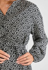 Schwarz-weiß gemustertes Kleid mit langen Ärmeln, Taillenknoten und lockerer Passform. Der Stoff hat eine weiche Textur und einen subtilen Glanz.