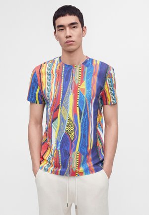 Ung mand iført en kortærmet flerfarvet striped og mønstret t-shirt, stående med hænderne i lommerne mod en ensfarvet baggrund.