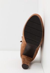 Brązowy zamszowy botek za kostkę z czarną gumową podeszwą; cechuje się teksturowanym gripem, zakrzywionym kształtem i delikatnym obcasem.