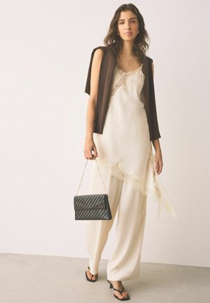 Femeie purtând o rochie crem din dantelă și pantaloni largi, cu un pulover maro pe umeri, ținând o poșetă neagră cu lanț auriu.