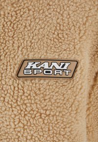 Kahvabeige fliiskangas, millel on tekstuur. Peal on keskelt tikitud must ja beeži "KANI SPORT" silt.