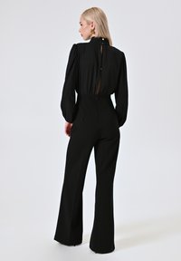 Zwarte jumpsuit met lange, pofmouwen, hoge hals en een keyhole rug. Uitlopende broekspijpen en gladde stof, voorzien van een discrete ritssluiting.
