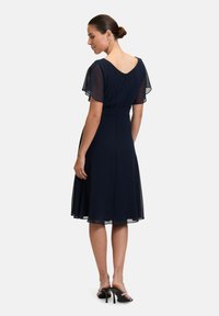Robe de marin avec des manches courtes en voile, taille cintrée et jupe évasée. L'arrière présente un décolleté subtil et une fermeture éclair. Portée avec des escarpins noirs.