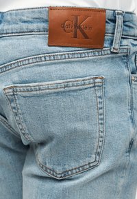 Φωτεινά γαλάζια τζιν από denim με ένα καφέ δερμάτινο λογότυπο Calvin Klein πάνω από την πίσω τσέπη, φορεμένα με λευκή μπλούζα.