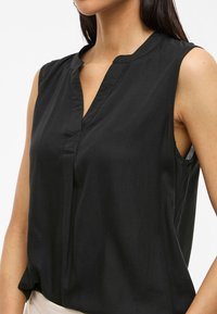 Musta ilmanhihaista puseroa, jossa on V-pääntie, pehmeä kangas ja hieman löysä istuvuus. Huomaamaton saumarakenne etuosassa.