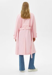 Long pink trench coat with a tied waist, open sleeves, and a back slit, paired with flared blue jeans and light-colored shoes.
