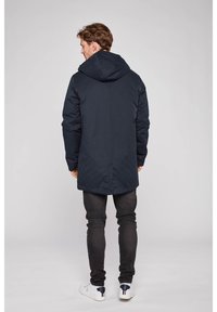 Navy zateplená bunda s kapucí, dlouhého střihu a hladkou texturou. Nošena s černými džíny a bílými teniskami s námořnickými akcenty.