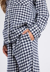 Vrouw in een navy- en witgeruite langemouwen blouse met bijpassende broek, met hand in de broekzak.