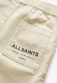 Beige tygshorts med resårmidja och en bakficka med logotypen "ALLSAINTS" samt japansk text i svart tryck.
