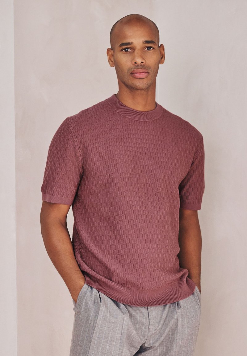 Homme les mains dans les poches, portant un pull à manches courtes texturé mauve et un pantalon gris clair à fines rayures sur fond uni.