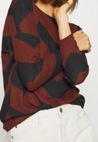 Suéter con patrón de zigzag en rojo y negro, de mangas largas y corte relajado, combinado con pantalones blancos que tienen un cierre de botón plateado.